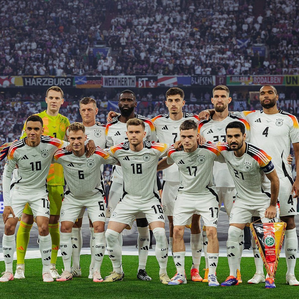 The German national football team poses together on the field in white jerseys with rainbow-striped sleeves before a match, with eleven players standing in two rows and a trophy placed in front of them.