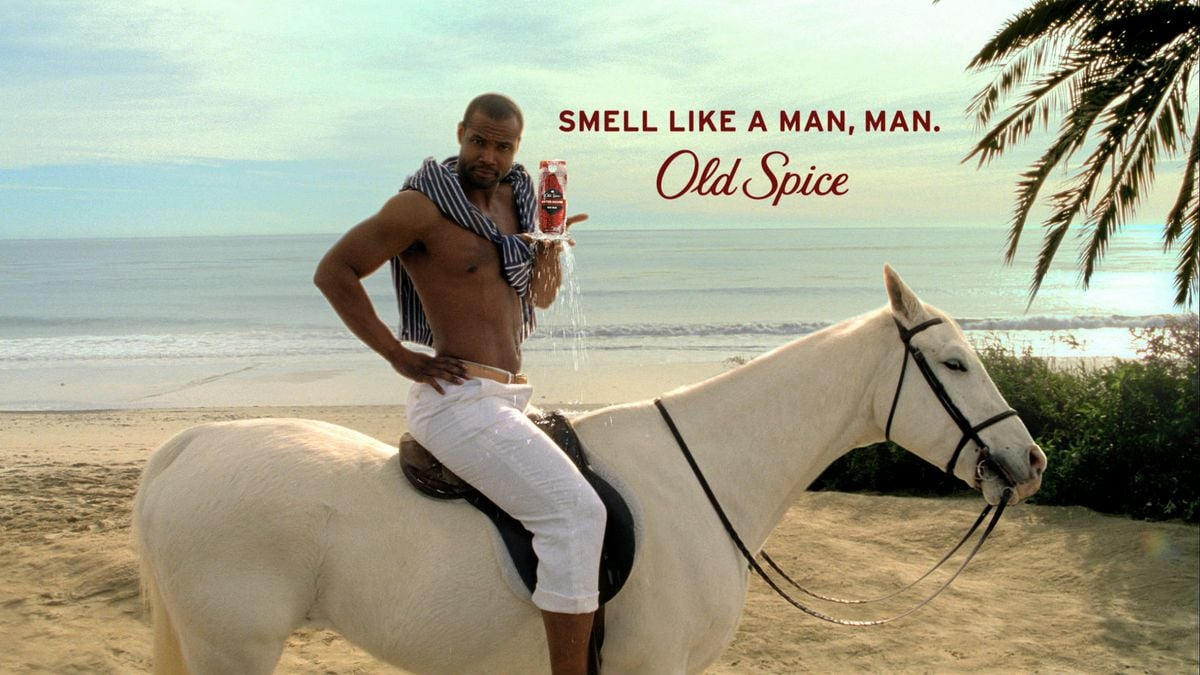 A shirtless man in white pants sits confidently on a white horse on a sandy beach, holding an Old Spice product. Text reads, "Smell like a man, man. Old Spice." Ocean and palm trees are in the background.