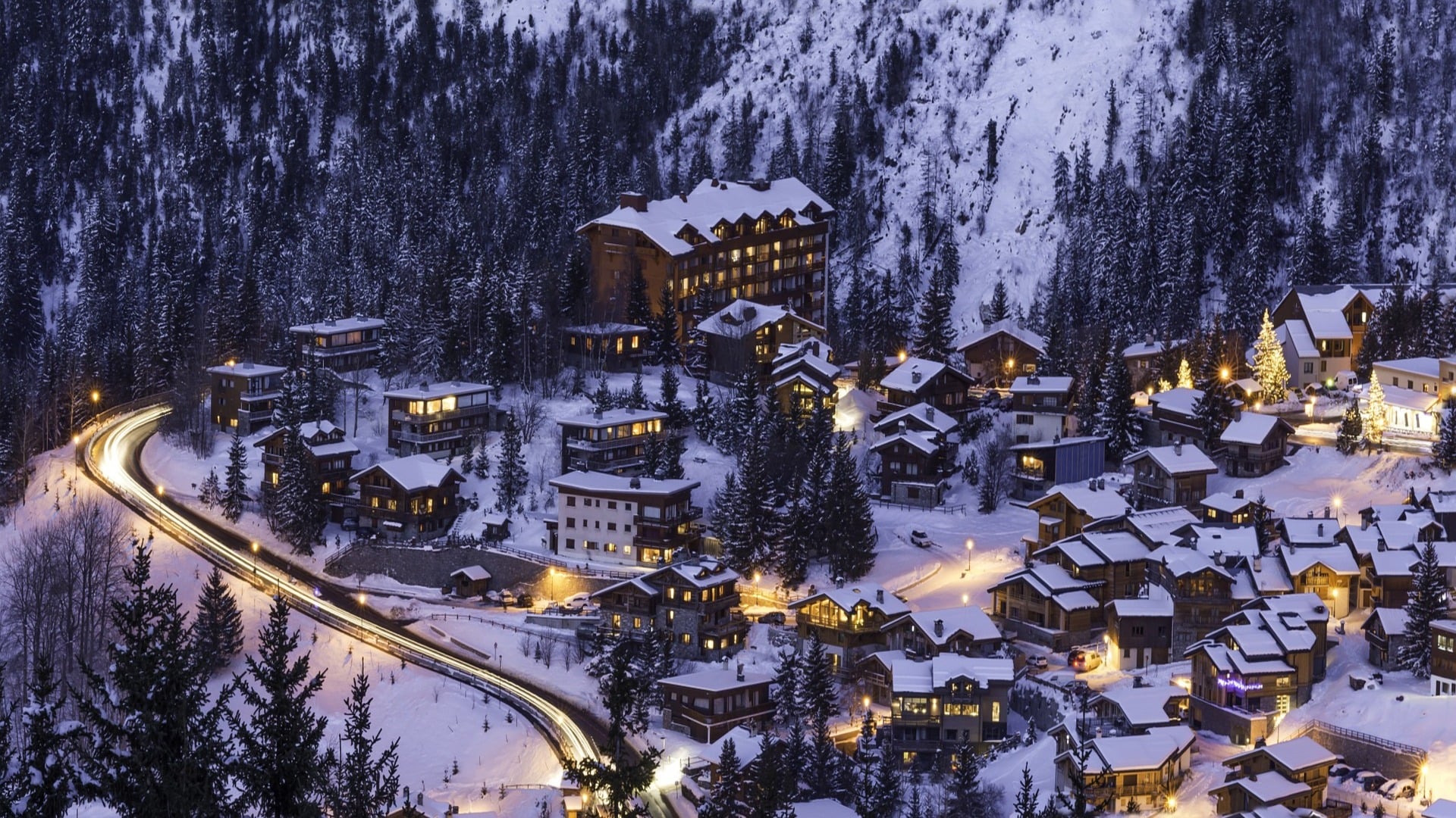 A snowy mountain village at dusk, with warmly lit chalets nestled among pine trees. A winding road with car light trails passes through the scene, and forested hills rise in the background.