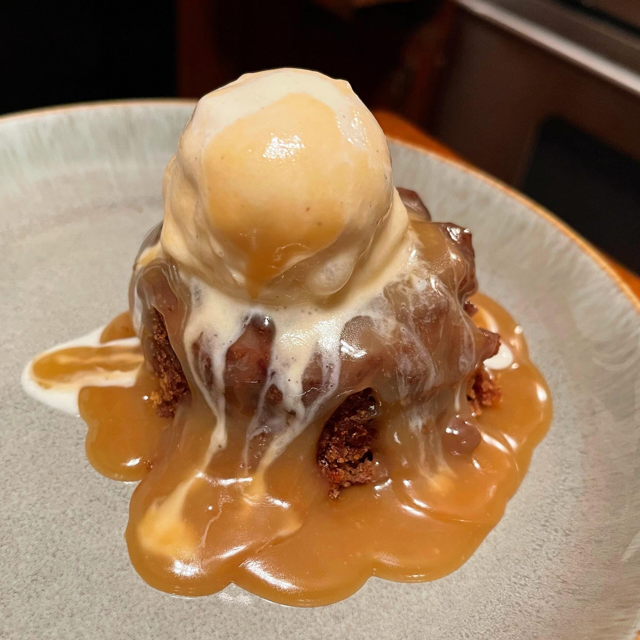 A scoop of vanilla ice cream melting over a warm cakey dessert, covered in rich caramel sauce, served on a light-colored plate.