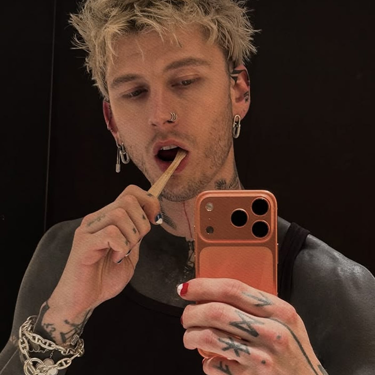 A person with blond hair, facial piercings, and tattoos takes a mirror selfie with a phone, holding a wooden stick in their mouth. They wear a black tank top, silver jewelry, and have painted nails.