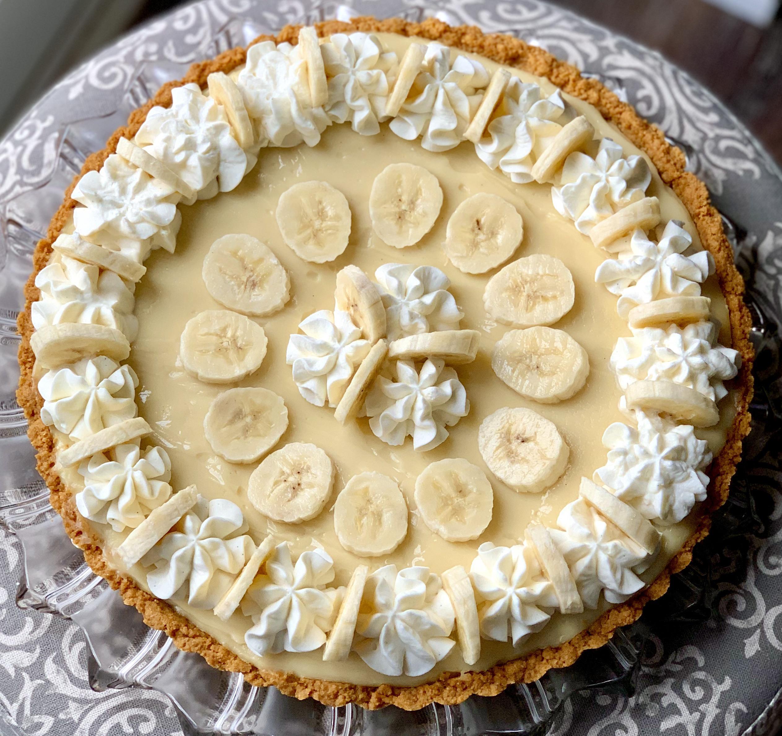 A banana cream pie with a graham cracker crust, topped with banana slices and decorative swirls of whipped cream arranged in concentric circles. The pie is displayed on a glass cake stand.