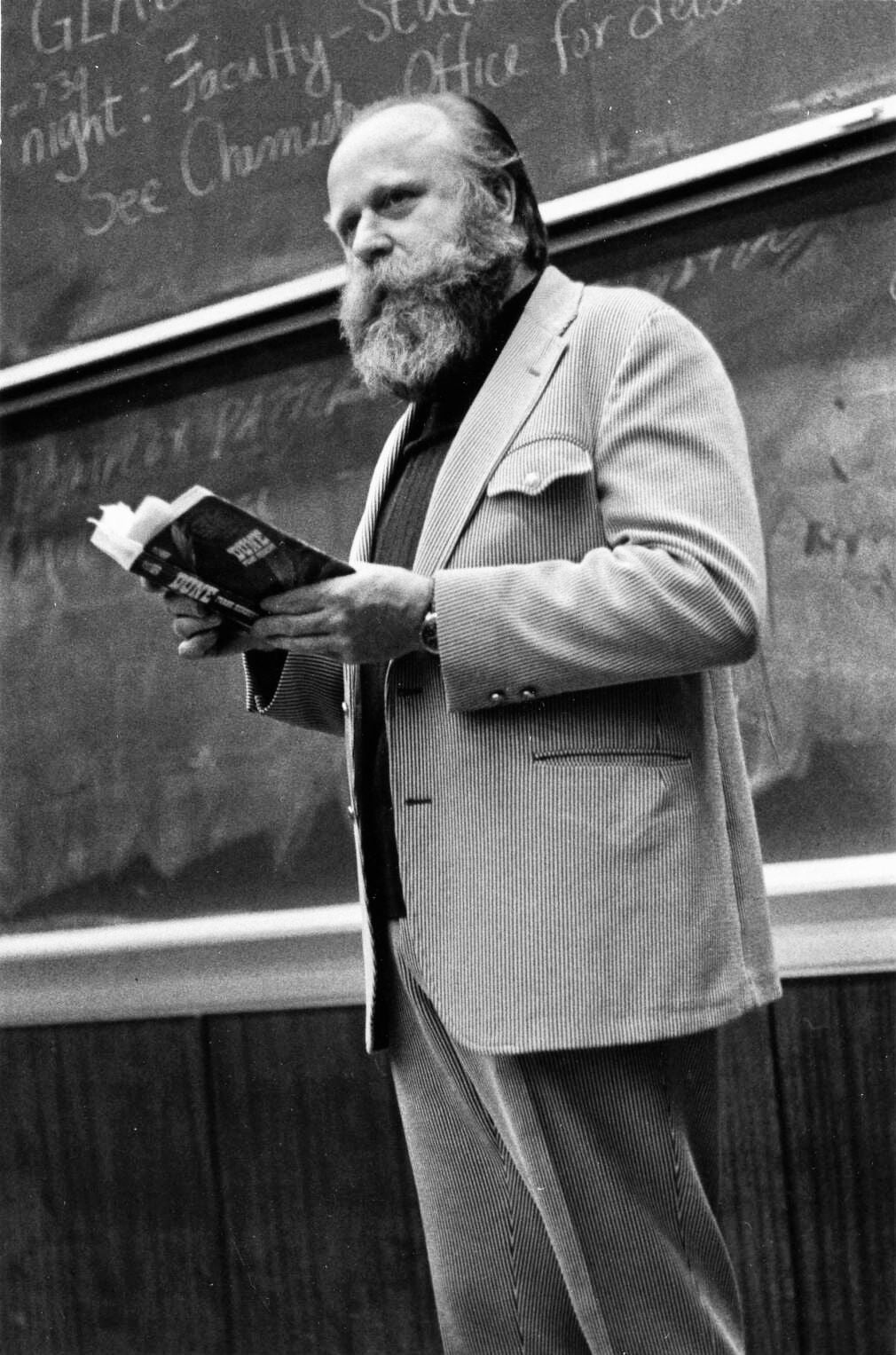 A man with a beard, wearing a striped suit, stands in front of a chalkboard holding an open book, appearing to lecture or speak in a classroom setting.