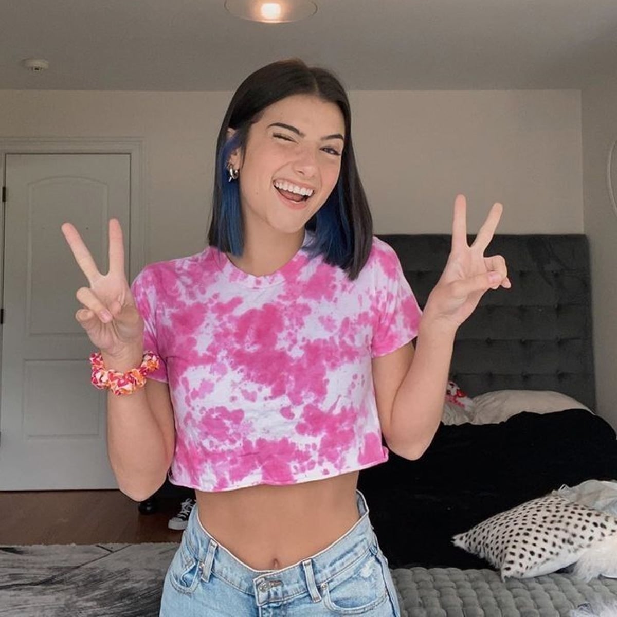 A young woman with dark hair and blue tips stands in a bedroom, smiling and making peace signs with both hands. She wears a pink tie-dye crop top and blue jeans. The bed and pillows are visible in the background.