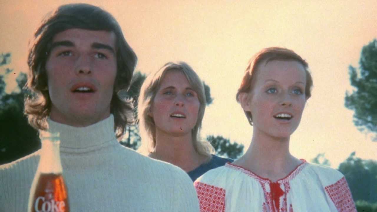 Three young people stand outdoors, looking ahead and singing. The person in the front left holds a glass bottle of Coca-Cola. All are bathed in warm sunlight, with trees and a bright sky in the background.