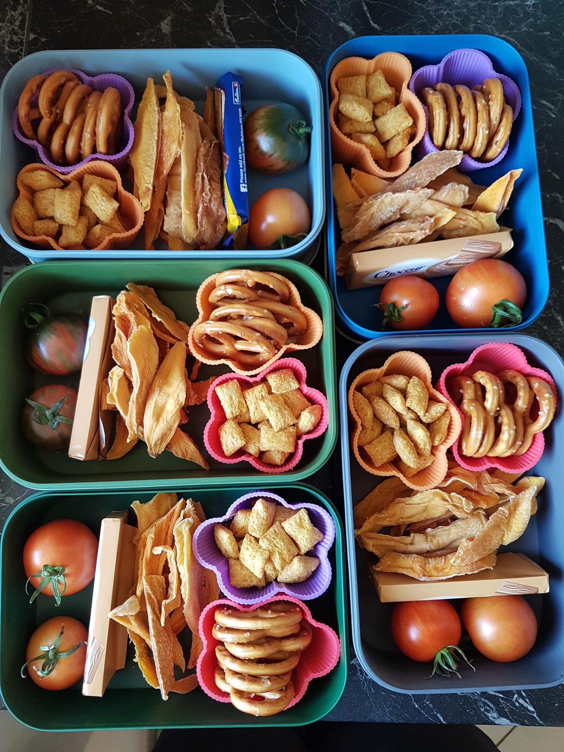 Five colorful lunchboxes filled with tomatoes, dried mango slices, pretzels, square breakfast cereal pieces, and a chocolate wafer bar, arranged neatly in silicone cups on a dark surface.