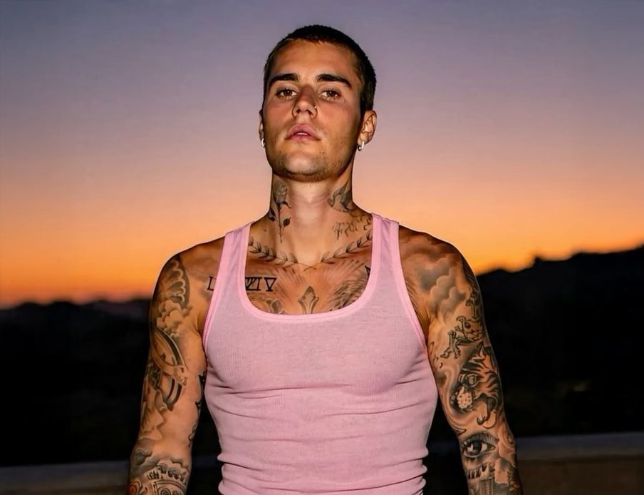 A young man with short dark hair, many tattoos, and wearing a light pink tank top stands outdoors at sunset with an orange and purple sky in the background.