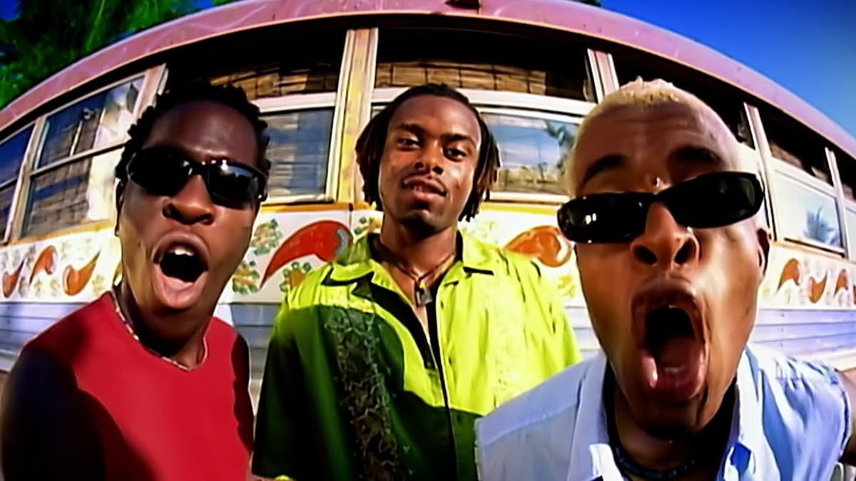 Three men wearing sunglasses stand in front of a colorful bus, two with mouths open wide, all looking at the camera on a sunny day. The man in the center wears a green shirt.