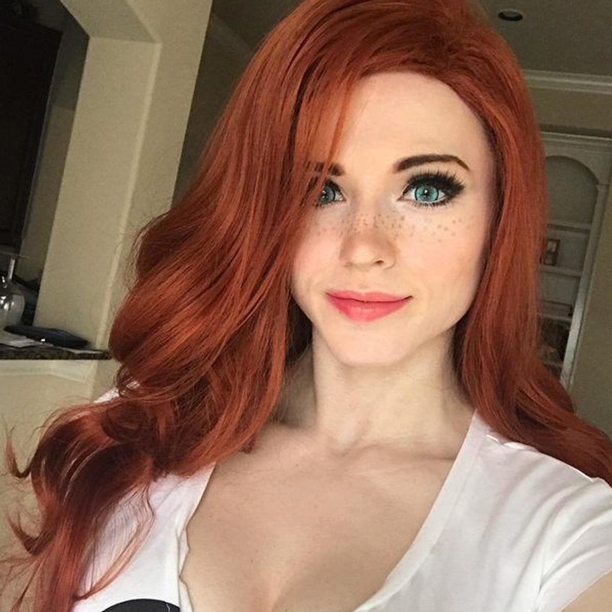 A woman with long, wavy red hair, blue eyes, and freckles smiles slightly while posing indoors. She is wearing a white top and is in a well-lit room with a neutral background.