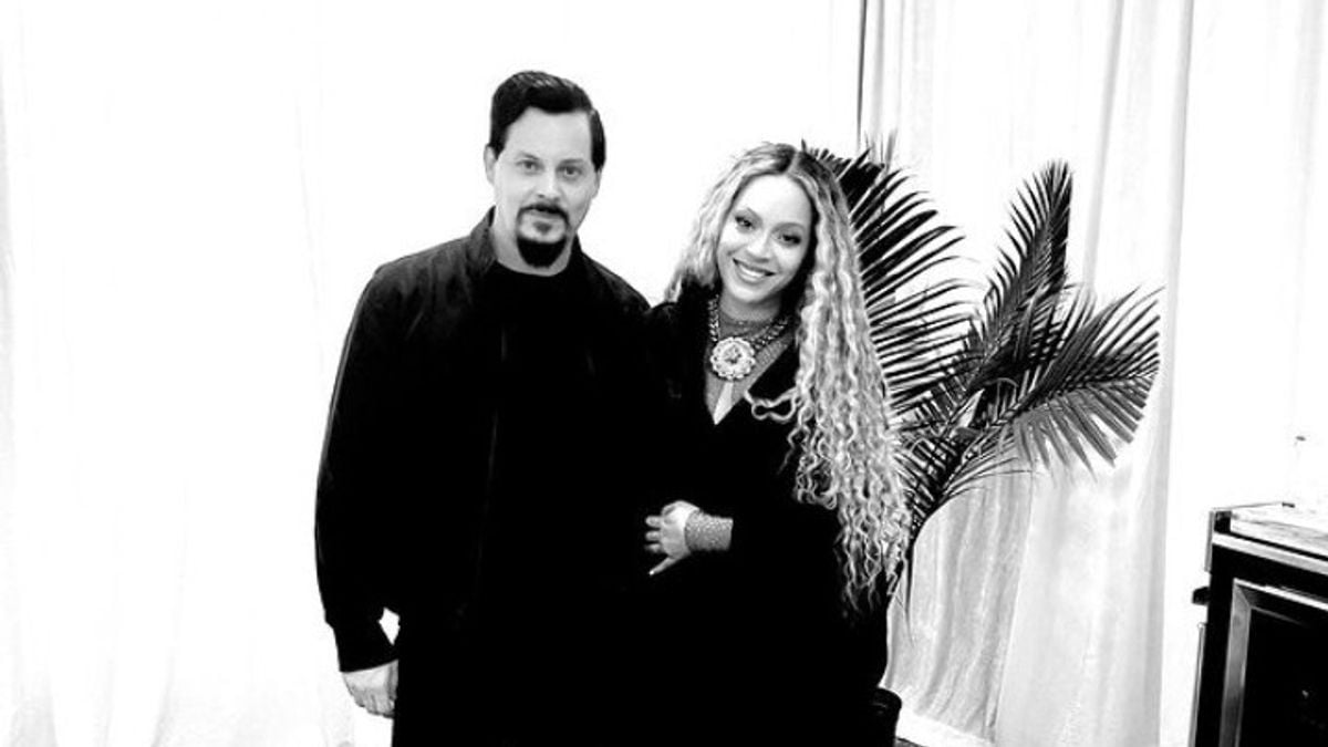 Two people stand together indoors in front of light-colored curtains and a potted plant. One has long curly hair, the other has short dark hair and a goatee. Both are dressed in dark clothing and smiling. The photo is in black and white.
