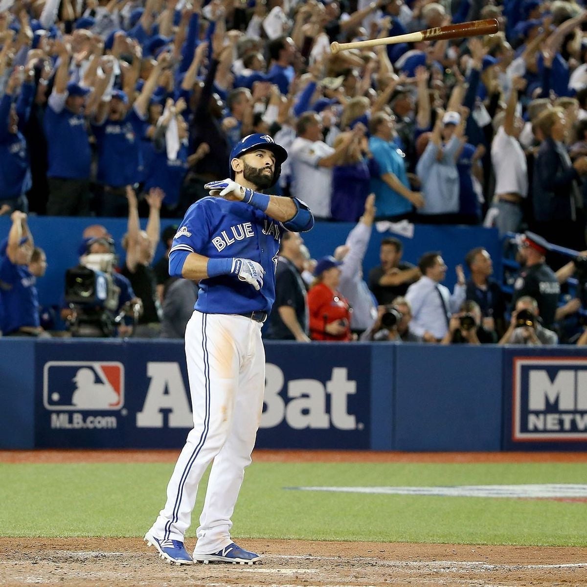A Toronto Blue Jays baseball player flips his bat dramatically after hitting, while a large crowd of excited fans in blue cheers in the background.