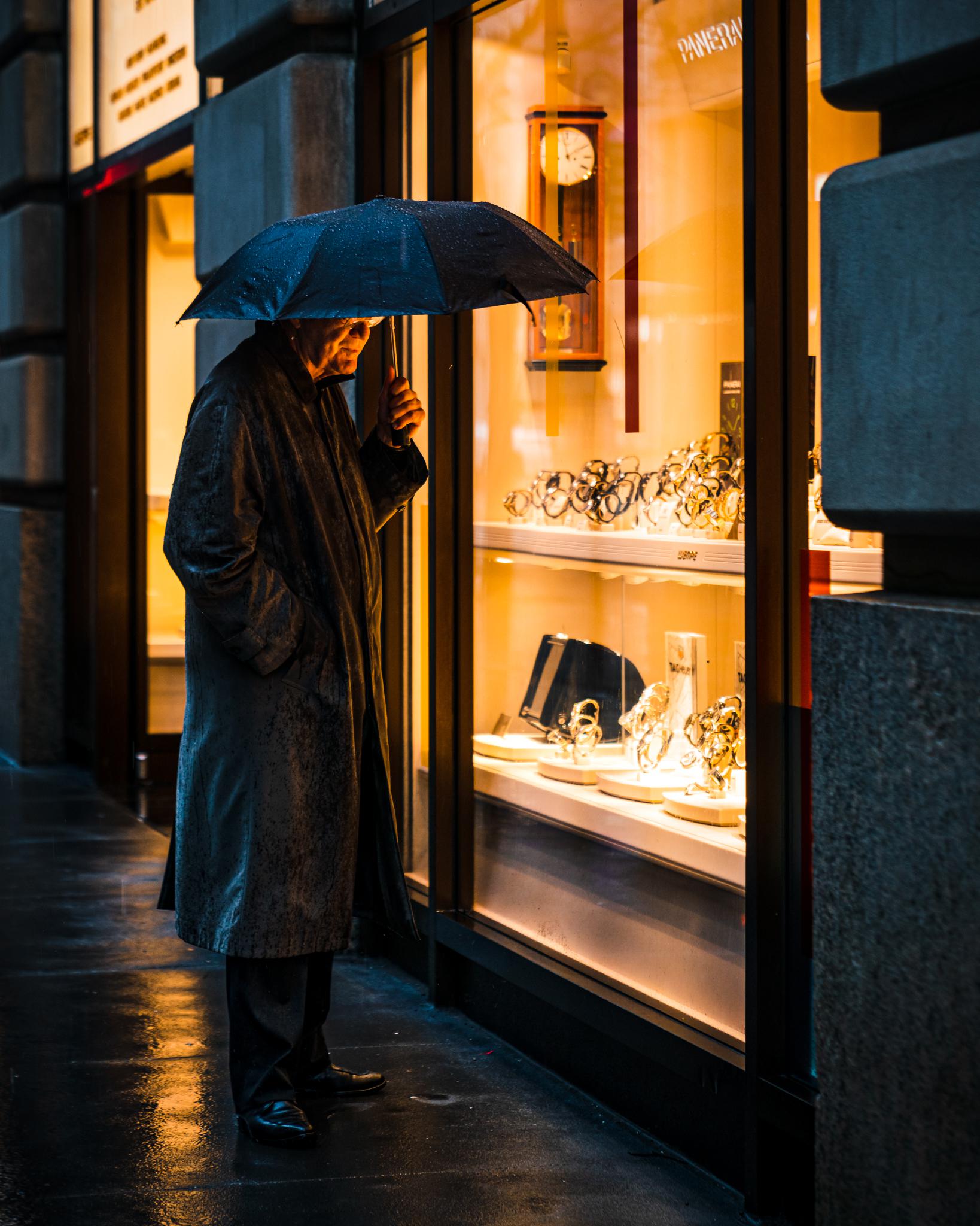 A person in a dark coat holding a black umbrella stands on a wet sidewalk, looking into a brightly lit shop window displaying luxury watches and clocks.