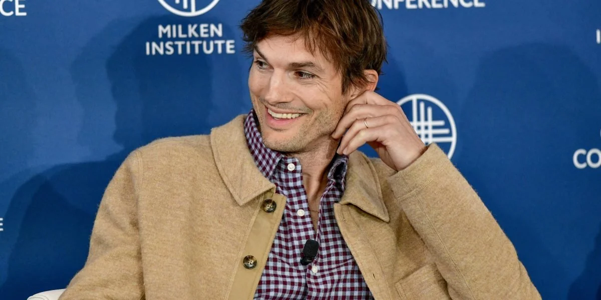 A man with brown hair in a tan jacket and checkered shirt sits smiling and touching his ear, in front of a blue backdrop with "Milken Institute" and conference graphics.