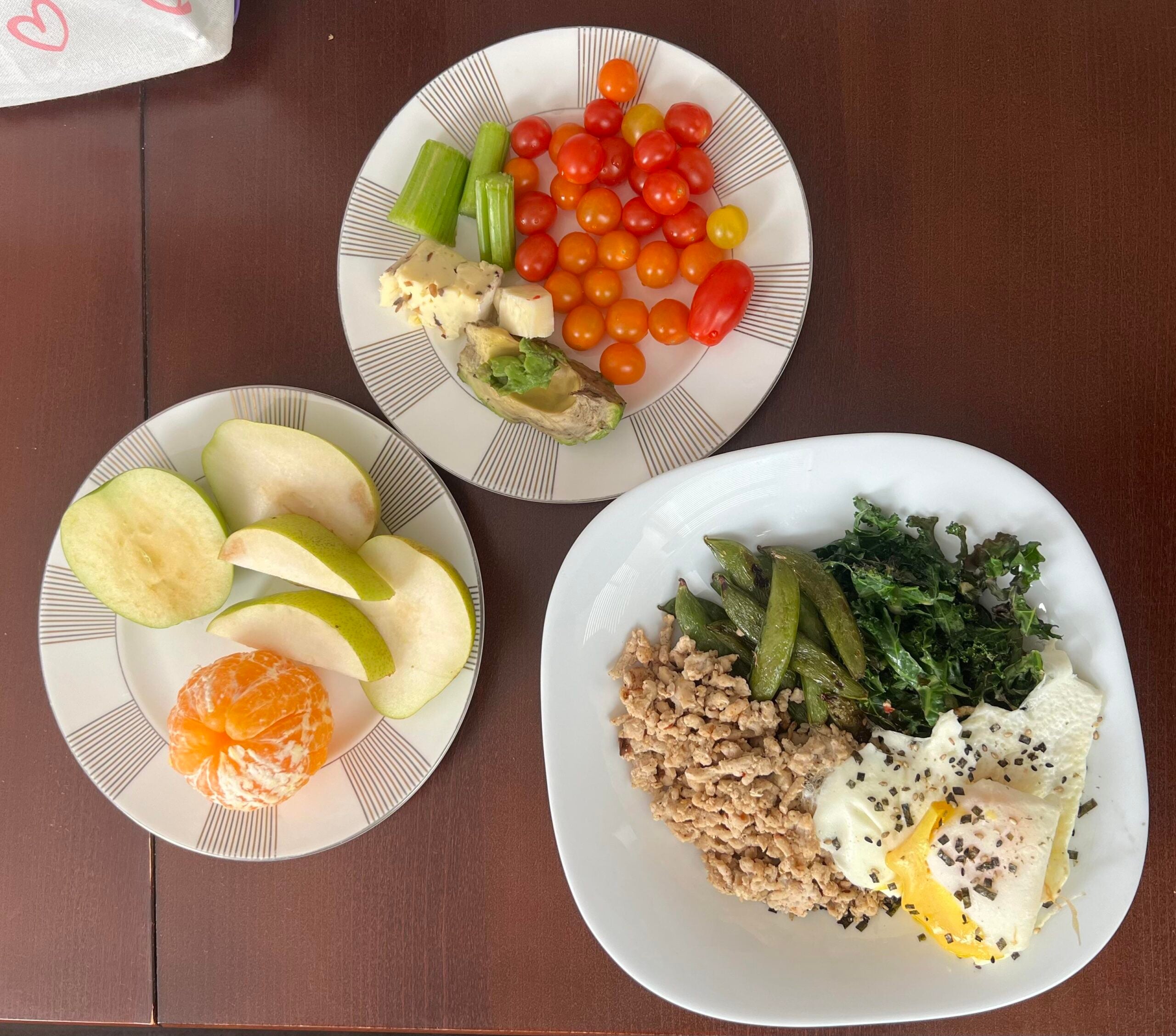 Three plates of food on a brown table: one with cherry tomatoes, celery, cheese, and avocado; one with apple slices, pear slices, and a peeled mandarin; and one with ground meat, greens, snap peas, and two fried eggs.