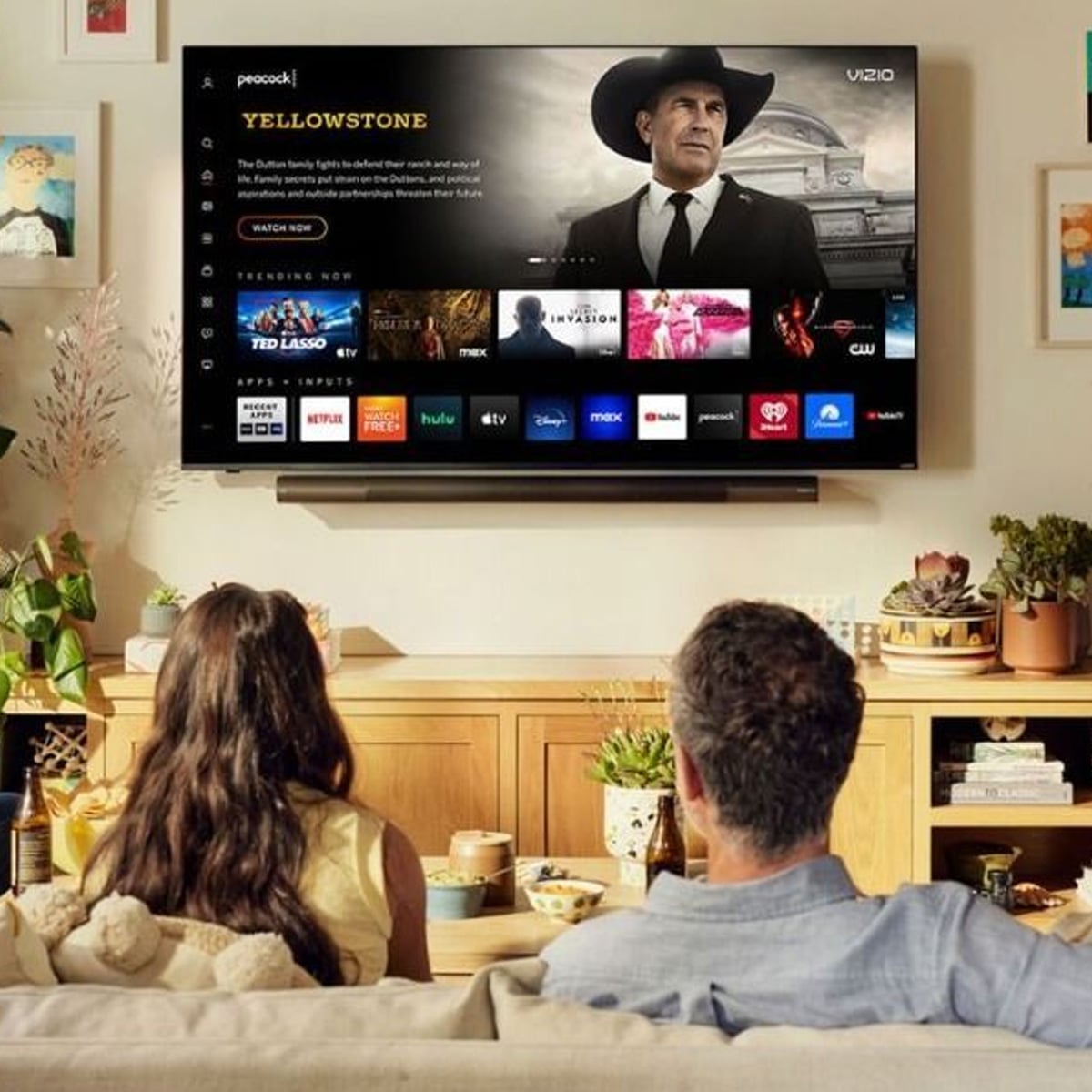 A couple sits on a couch in a cozy living room, watching a large wall-mounted TV displaying the streaming menu for "Yellowstone" and other shows. The room is decorated with plants and books.