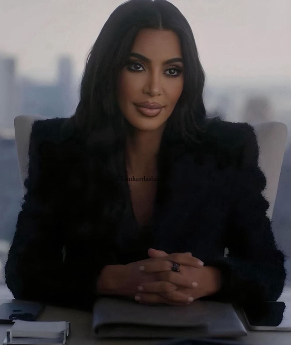 A woman with long dark hair and a black blazer sits at a desk with her hands clasped, looking forward. There are papers and a pen on the desk, and a blurred cityscape is visible in the background.