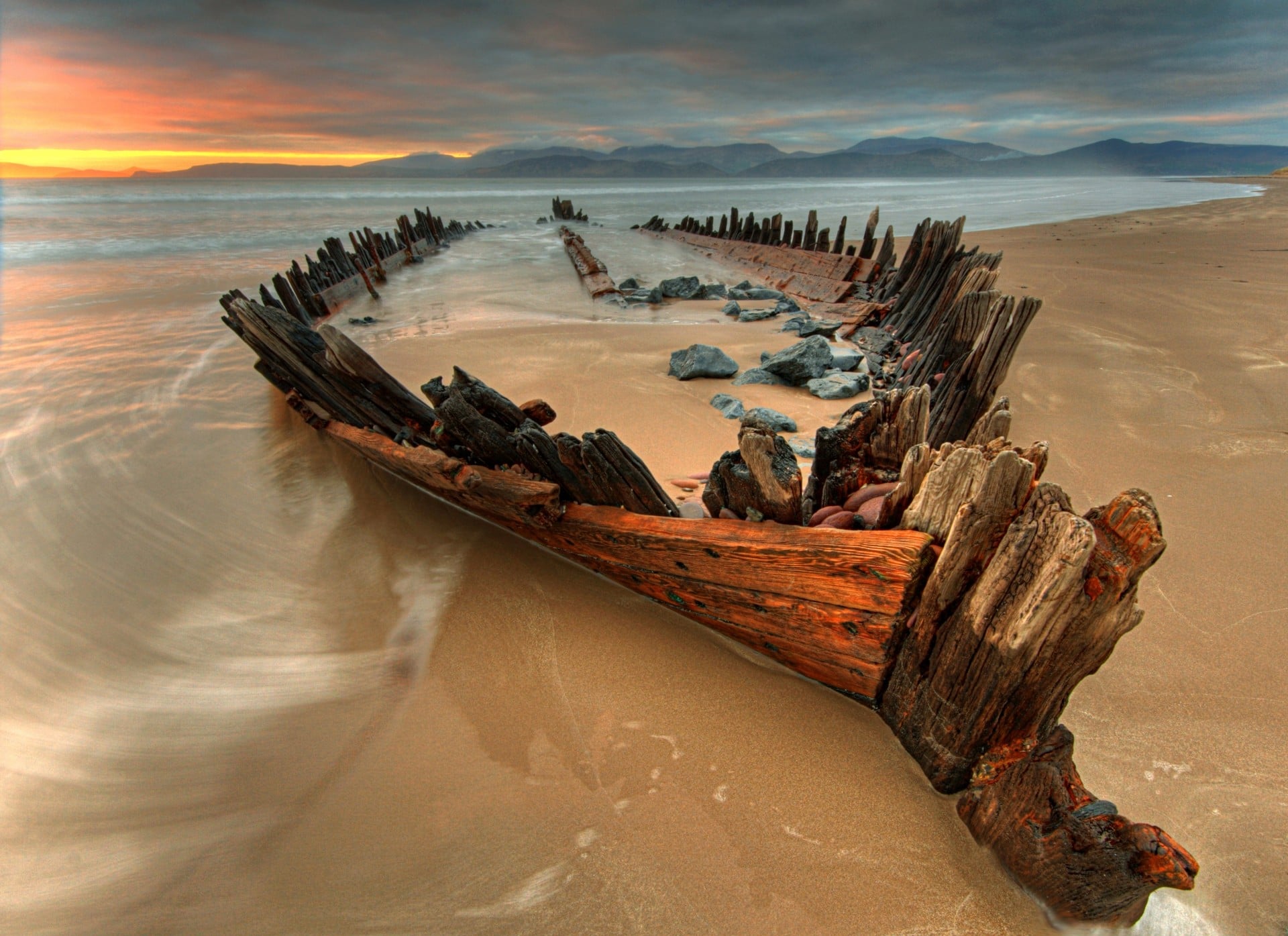 A weathered shipwreck with jagged wooden beams lies partially buried in the sand on a beach at sunset, with waves swirling around and mountains visible in the distance under a cloudy sky.
