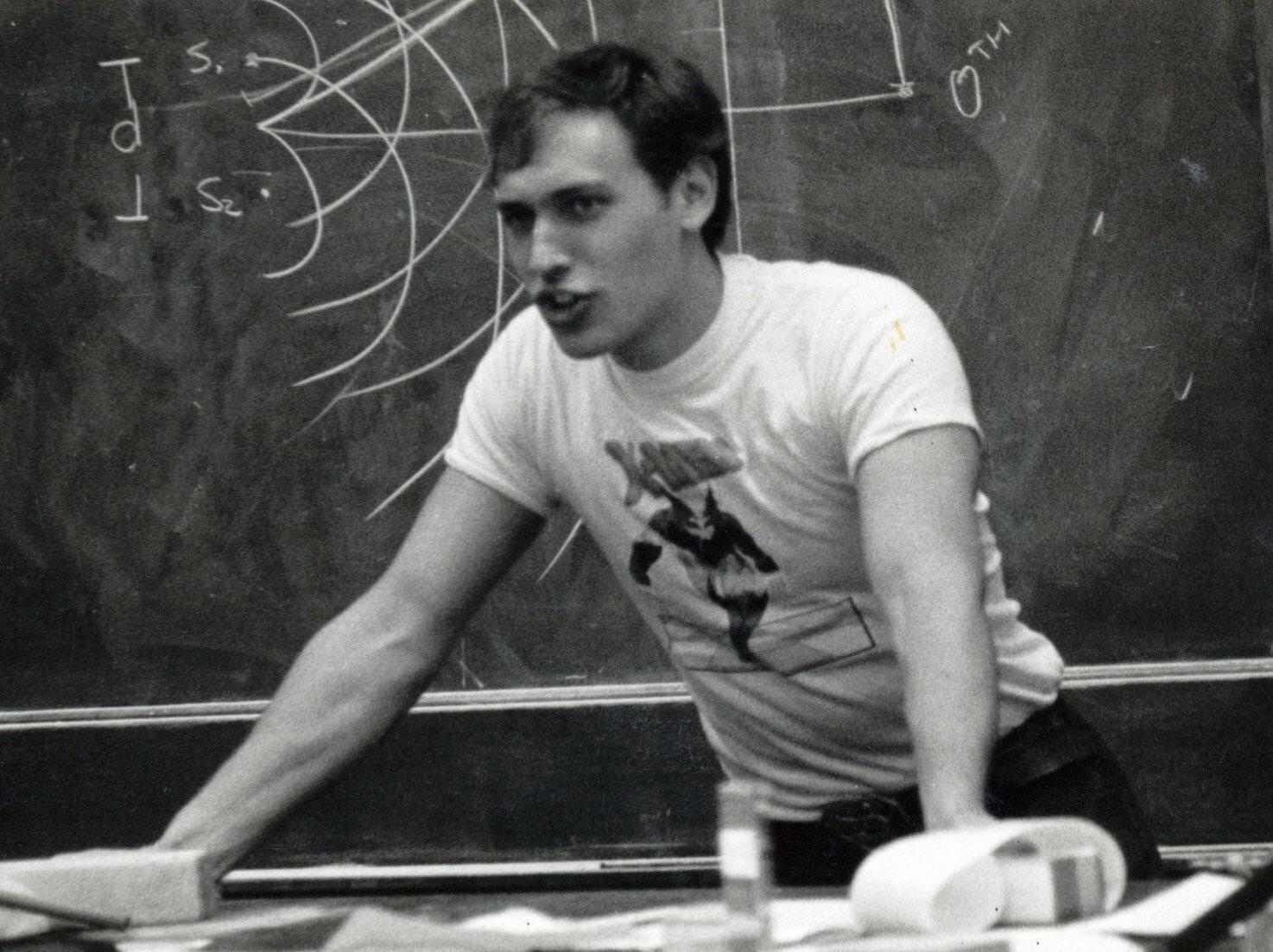 A young man in a graphic t-shirt leans over a desk, speaking animatedly in front of a chalkboard covered with diagrams and writing. Papers and objects are scattered on the desk.