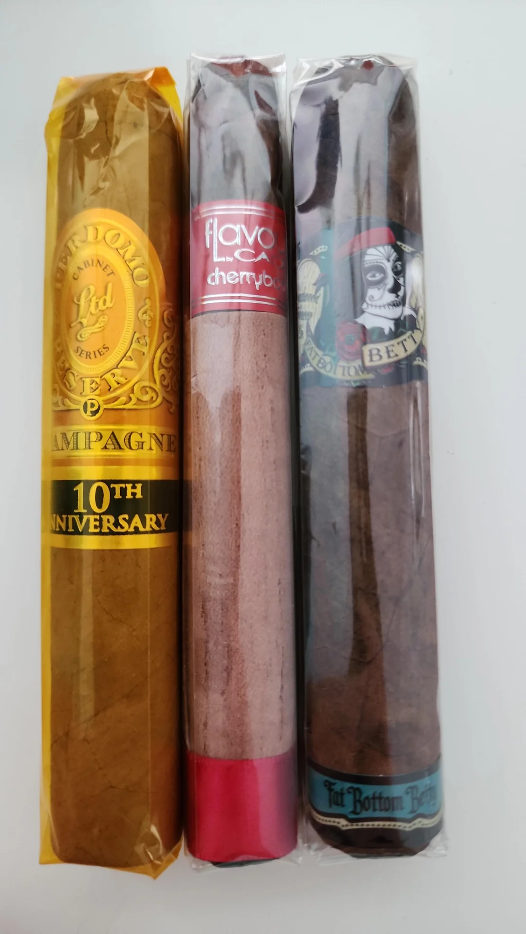 Three cigars in wrappers, each with a distinct label: one gold with "10th Anniversary," one red and silver with "cherry," and one dark with an illustrated face and the words "Fat Bottom Betty.