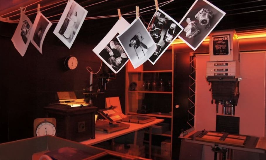 Photographs hang on a line to dry in a dimly lit darkroom with red lighting. The room contains photographic equipment, such as an enlarger, clocks, trays, and worktables.