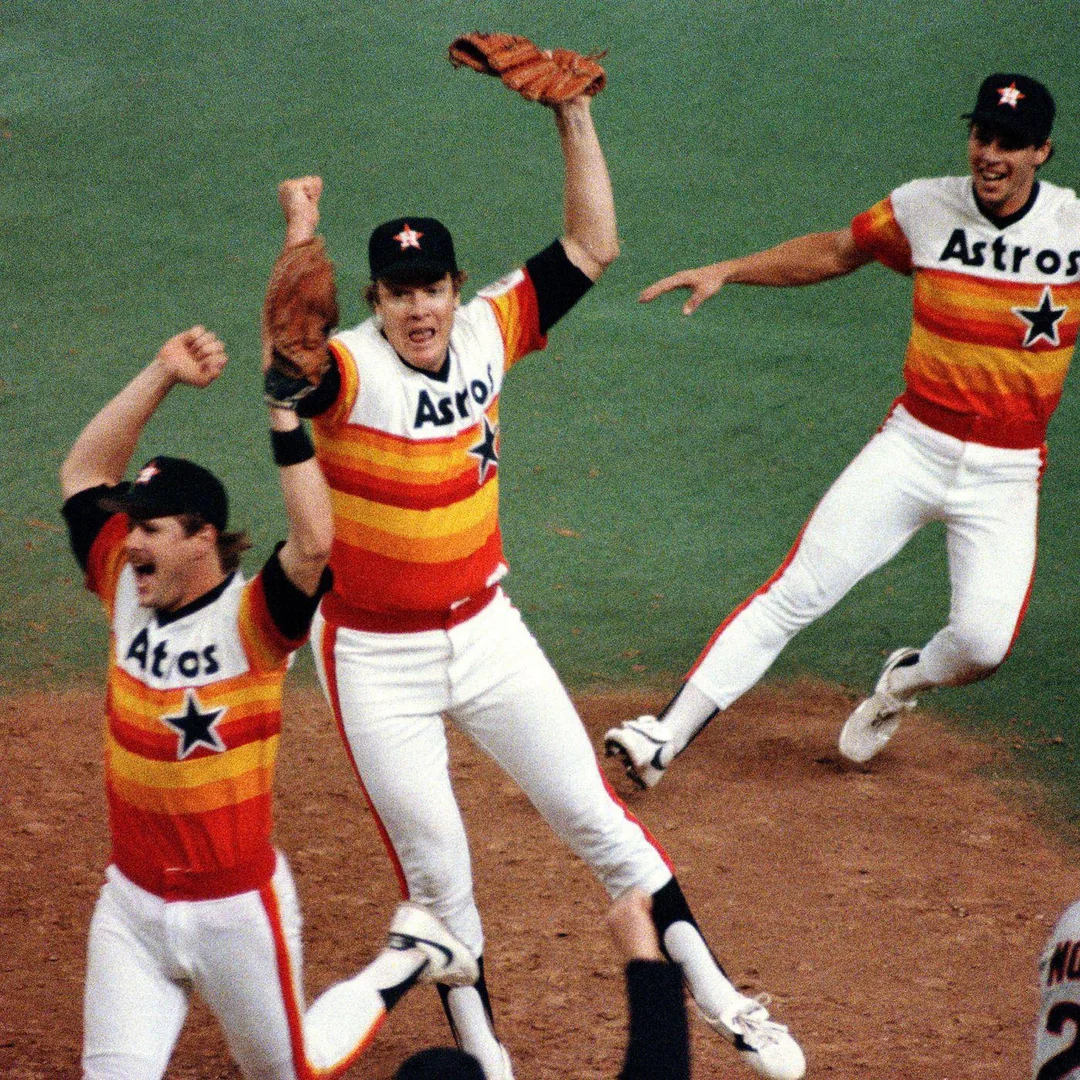 Three Houston Astros baseball players in colorful uniforms celebrate on the field, arms raised and smiling, after a game victory. The background shows part of the baseball diamond and green turf.