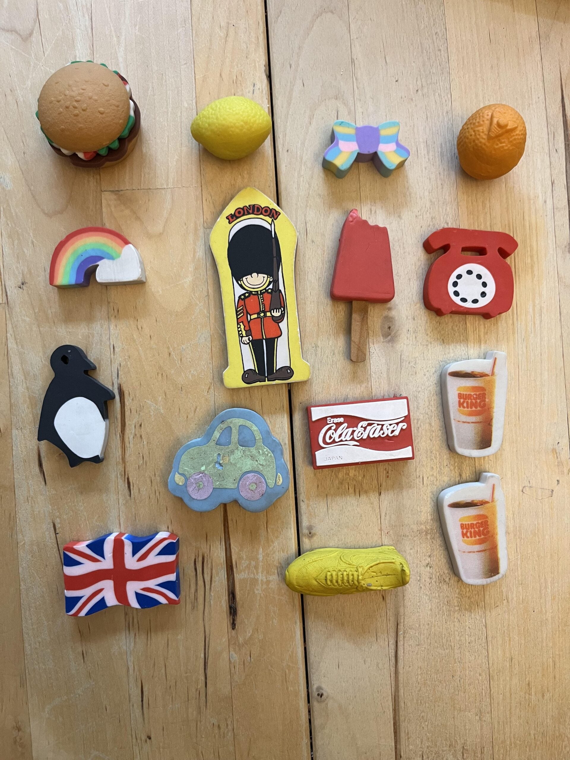 A collection of colorful erasers shaped like a burger, lemon, bow, orange, rainbow, British guard, popsicle, rotary phone, penguin, car, Coca-Cola sign, Union Jack, yellow sneaker, and two drinks on a wooden surface.
