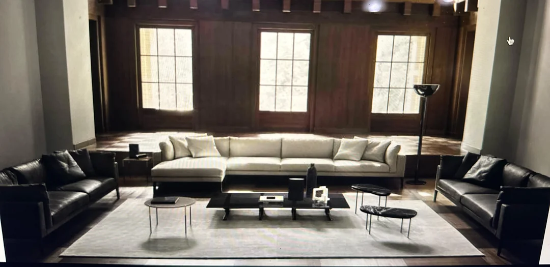 A spacious living room with dark wood paneling, large windows, a light-colored sectional sofa, two black leather sofas, a white rug, and several modern coffee tables and side tables.