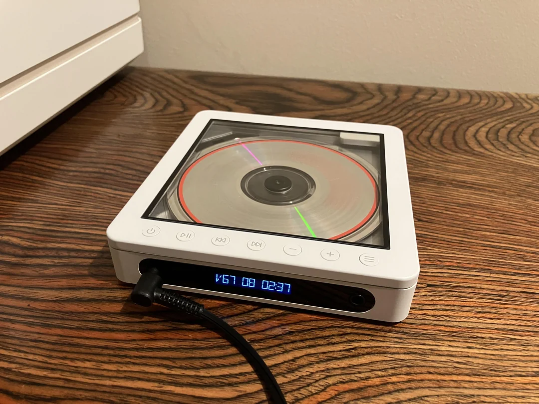 A white, modern CD player with a transparent lid is playing a disc on a wooden surface. A black cable is plugged in, and a digital display shows "67 08 02:37" in blue numbers.