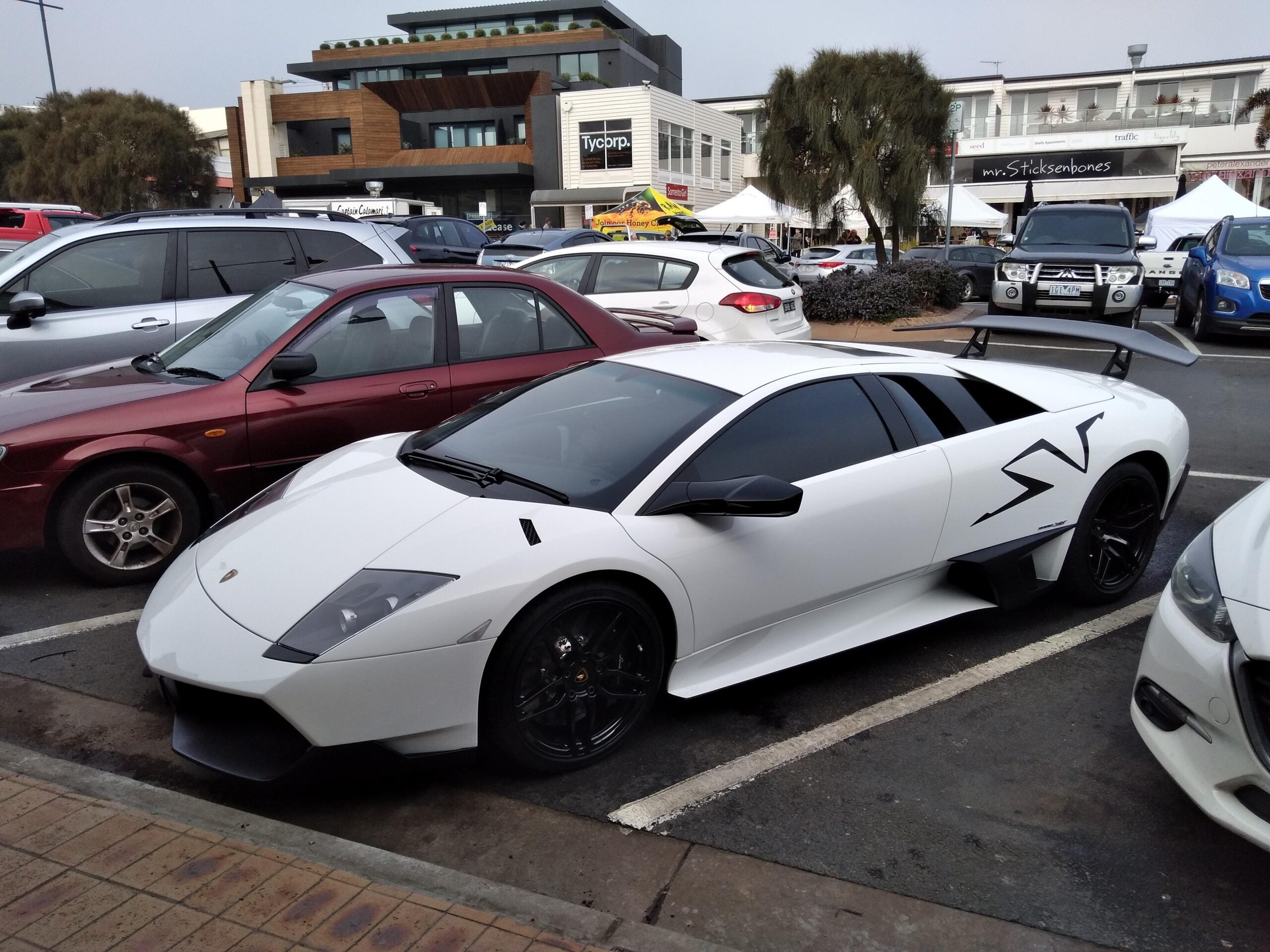 A white Lamborghini Murciélago SV with black accents is parked in a lot between other cars, with shops and trees visible in the background.