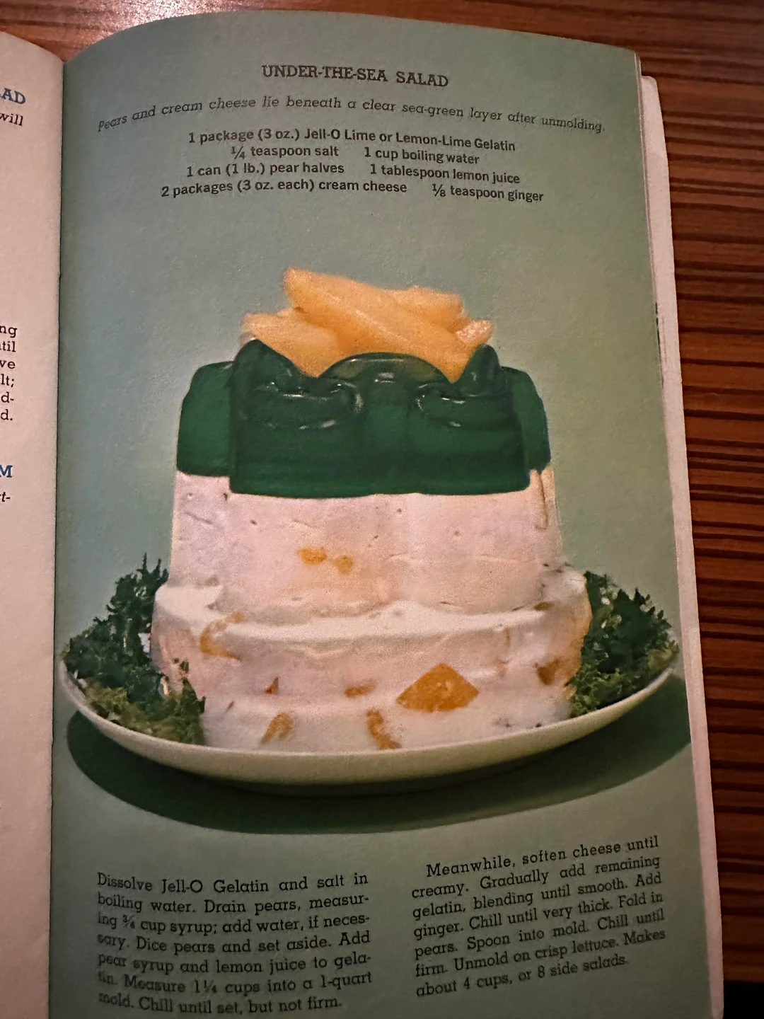 A vintage cookbook photo shows a molded salad with a green gelatin top layer, garnished with pear slices and surrounded by parsley. The bottom creamy layer contains chunks of fruit, and the dish sits on a white plate.