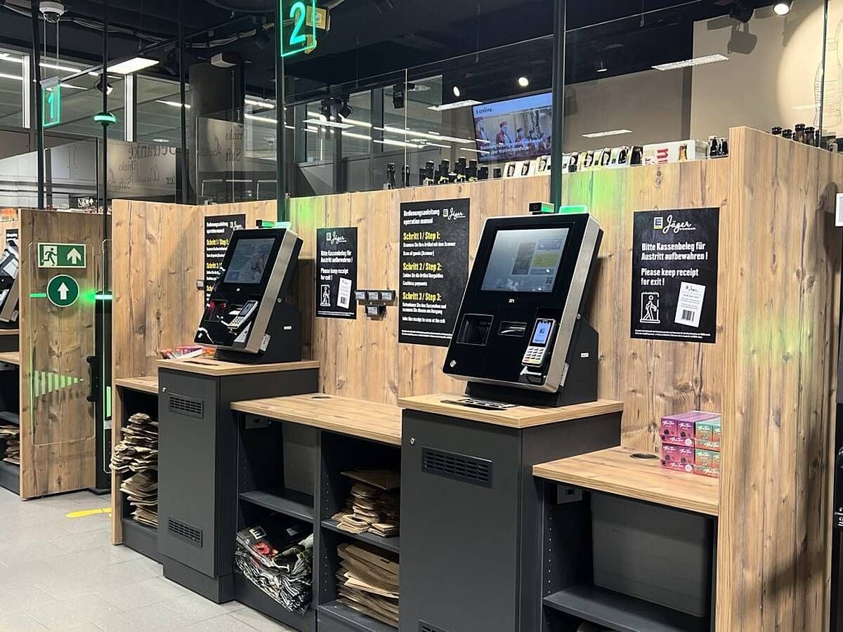 Self-checkout kiosks with touchscreens and card readers in a modern store, wooden counters below, and empty bag holders. Shelves with small items are visible on the right. Green exit signs hang above.