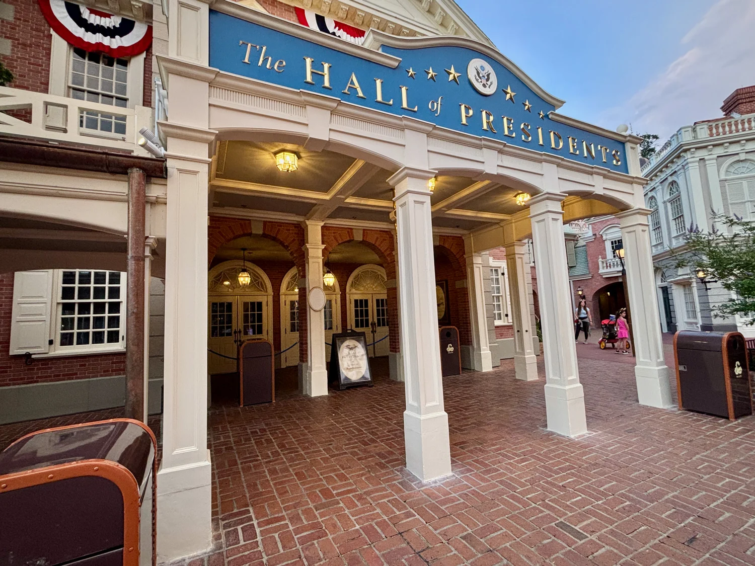 Entrance to The Hall of Presidents attraction, featuring a blue sign with gold letters, white columns, brick flooring, and early American-style architecture at a theme park.