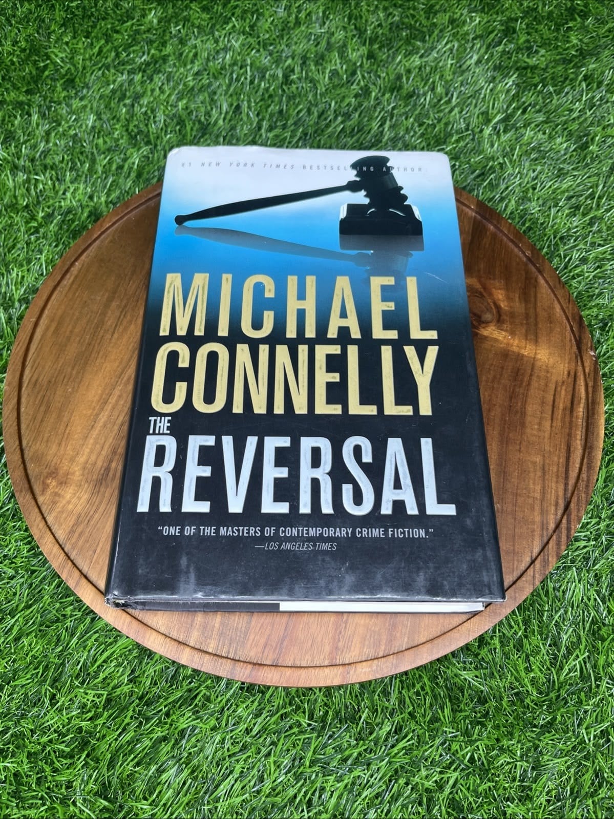 A hardcover copy of "The Reversal" by Michael Connelly is displayed on a round wooden board, placed on a background of green artificial grass.