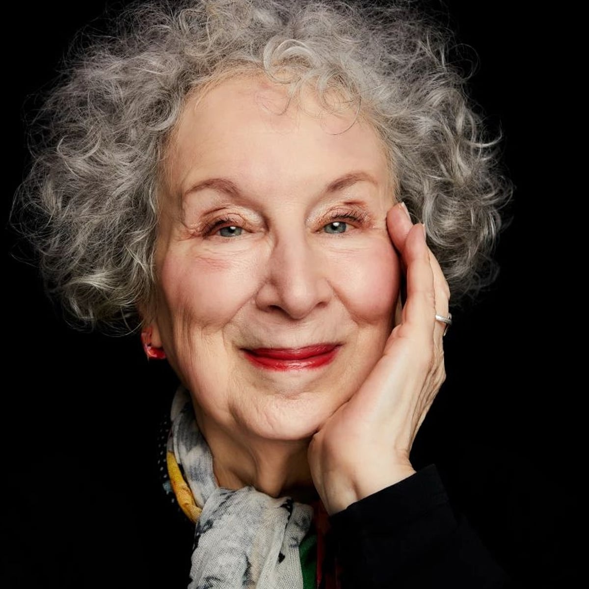An elderly woman with curly gray hair smiles warmly, resting her face on her hand. She wears a patterned scarf and black top, with a dark background highlighting her features.