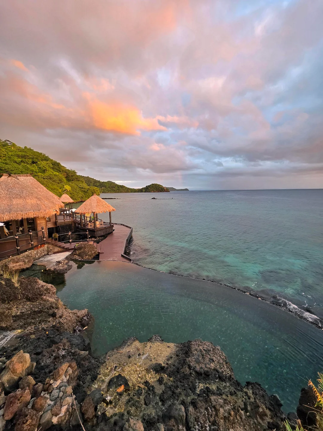 Thatched-roof huts stand on a wooden deck beside a calm, turquoise sea at sunset, with rocky shorelines and lush green hills under a colorful, cloud-filled sky.