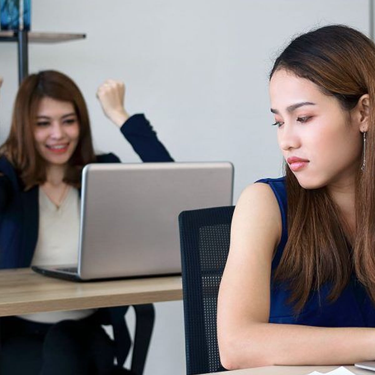 Two women at desks; one in the foreground looks sad and discouraged, while the other in the background cheers joyfully with raised fists in front of her laptop.