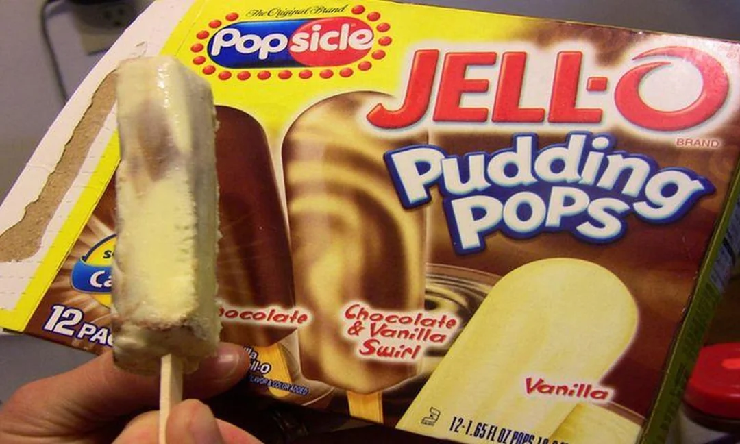 A hand holding a vanilla and chocolate swirl Jell-O Pudding Pop with a box of Popsicle brand Jell-O Pudding Pops in chocolate, swirl, and vanilla flavors in the background.