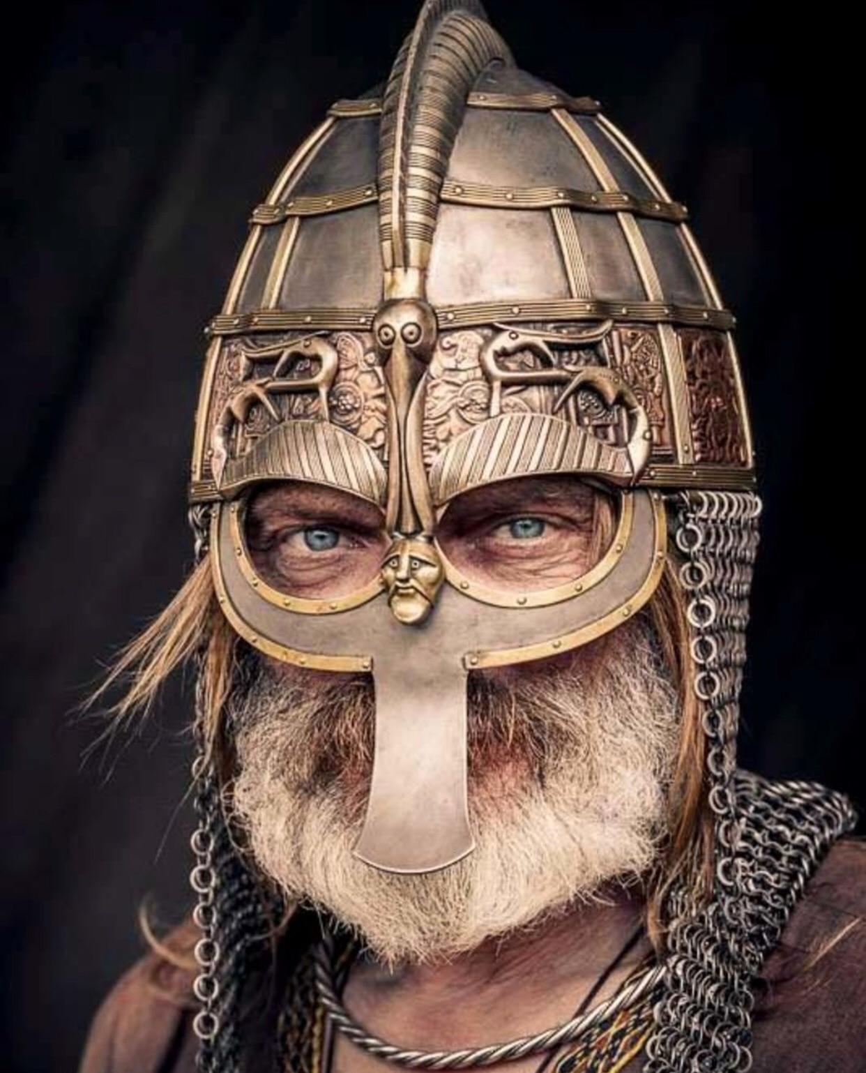A man with a white beard and intense blue eyes wears an ornate metal helmet and chainmail. The helmet features intricate carvings, a nose guard, and face details, evoking ancient Viking or medieval warrior armor.
