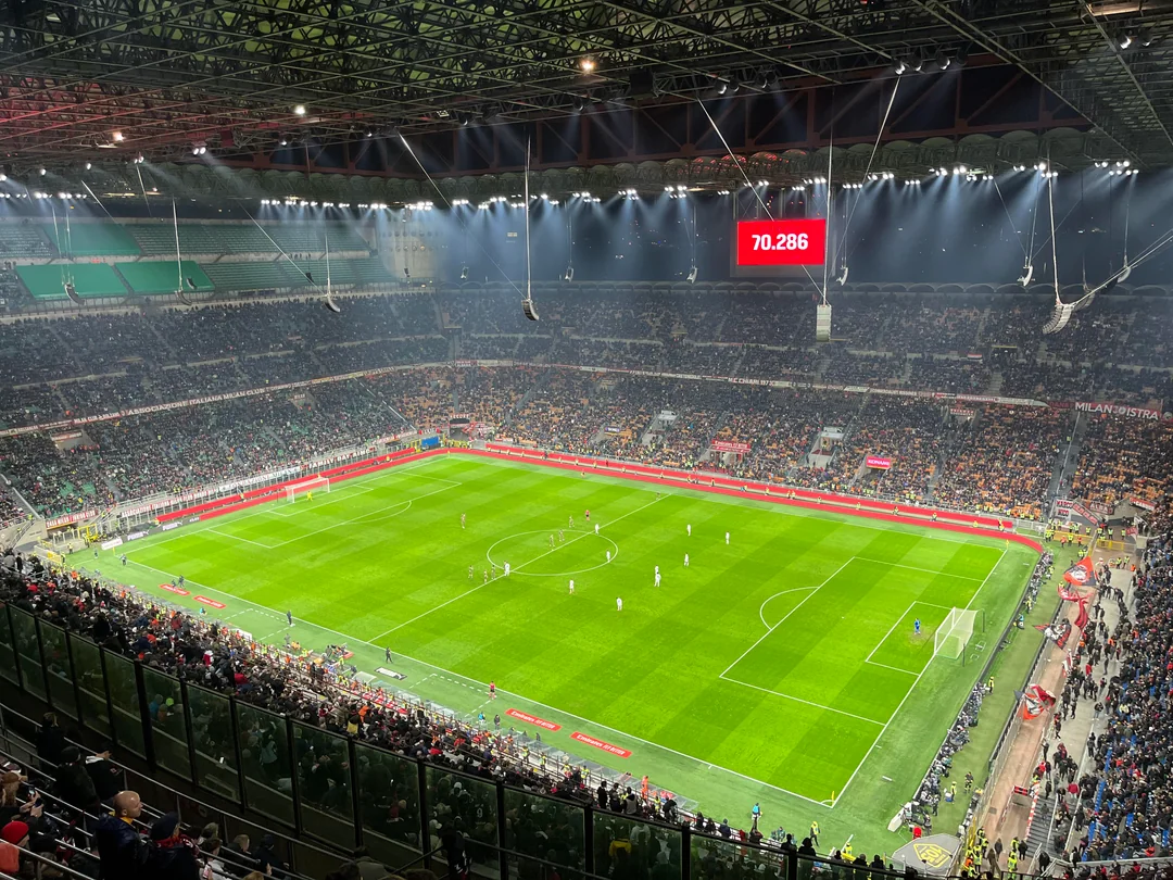 A large, brightly lit football stadium is filled with spectators watching a match. The scoreboard displays "70,286" above the field, and players are spread out across the green pitch.