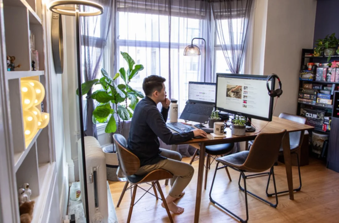 A person sits at a desk in a bright home office with large windows, working on a computer. The space features a houseplant, bookshelves, a headset, and a water bottle, creating a cozy and organized atmosphere.
