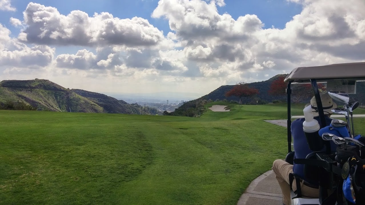 A scenic view of a golf course with lush green grass, rolling hills, and a cloudy sky. A golf cart with golf clubs is parked on a path in the foreground to the right.
