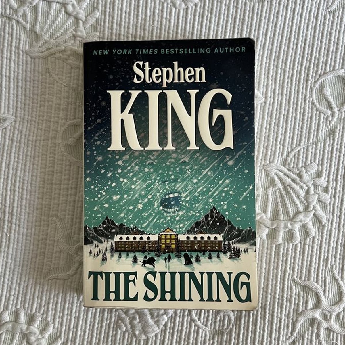 A copy of Stephen King's novel "The Shining" rests on a white textured surface. The cover shows a snowy scene with mountains, a large building, trees, and shadowy figures in the foreground.