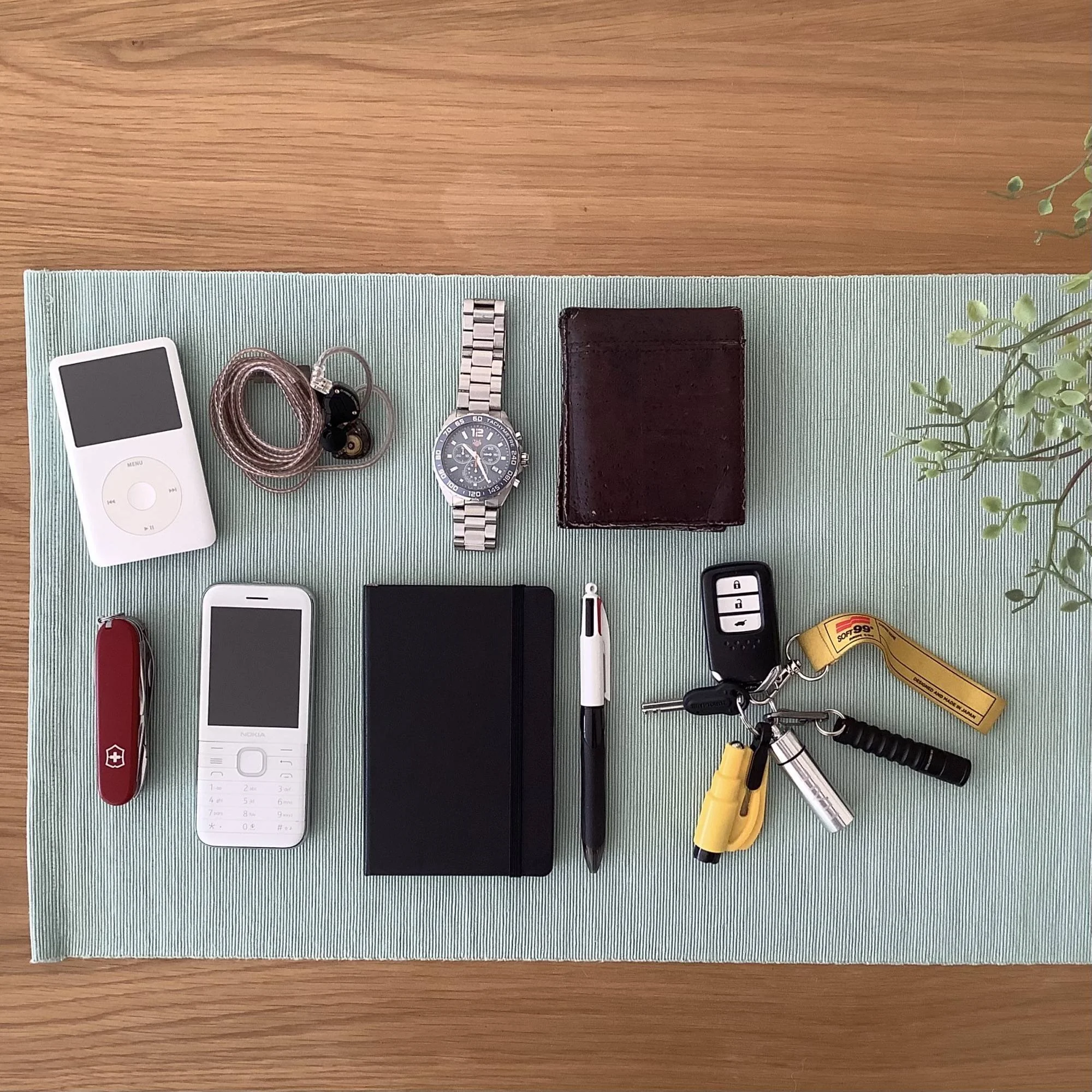 A flat lay of everyday items on a light placemat: an iPod, Swiss Army knife, mobile phone, watch, notebook, pen, leather wallet, and two sets of keys with various keychains, next to a plant.