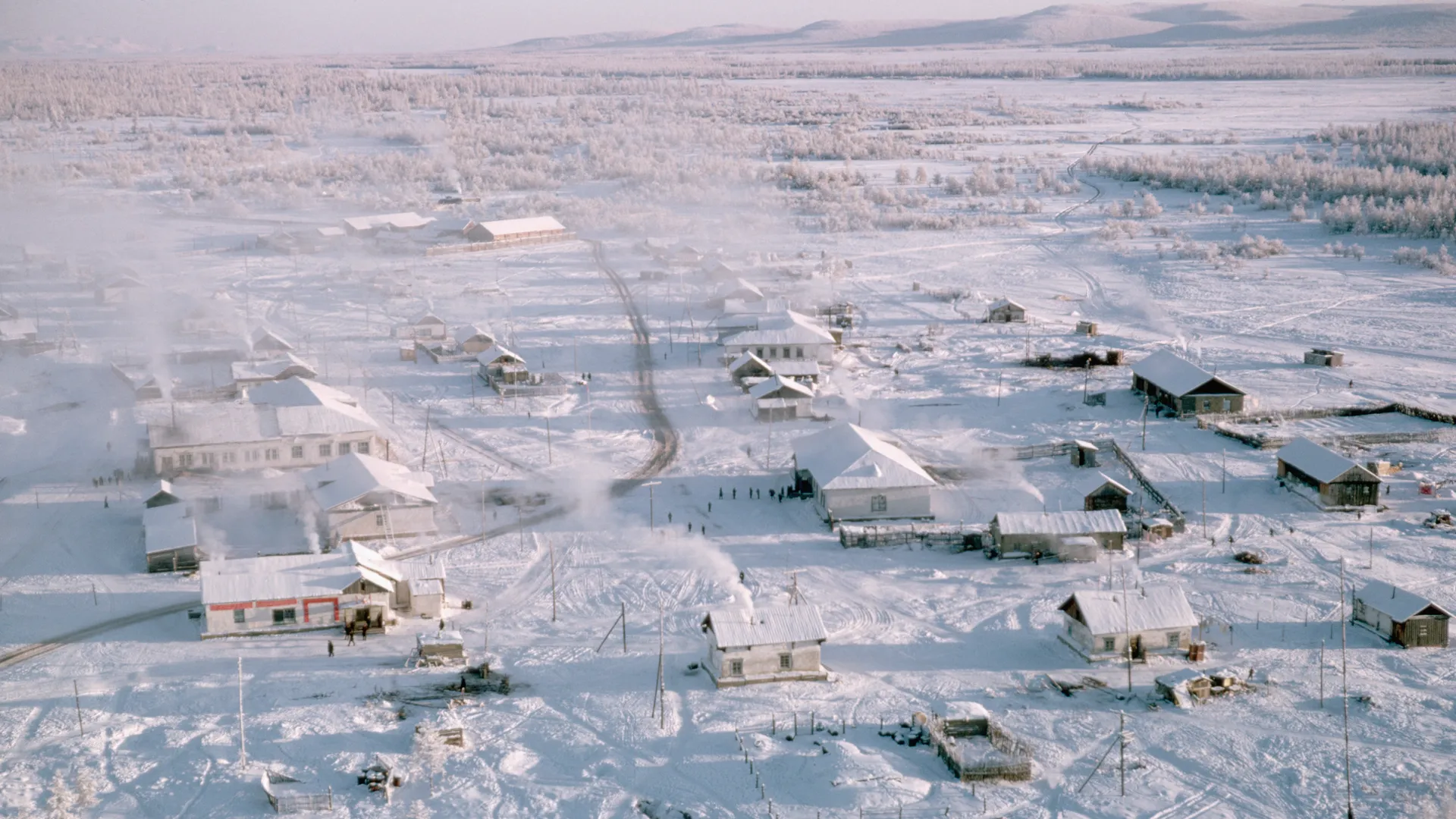 Aerial view of a rural village covered in snow, with scattered houses, roads, and smoke rising from chimneys. Surrounding landscape is frosty and expansive, with distant hills and leafless trees.