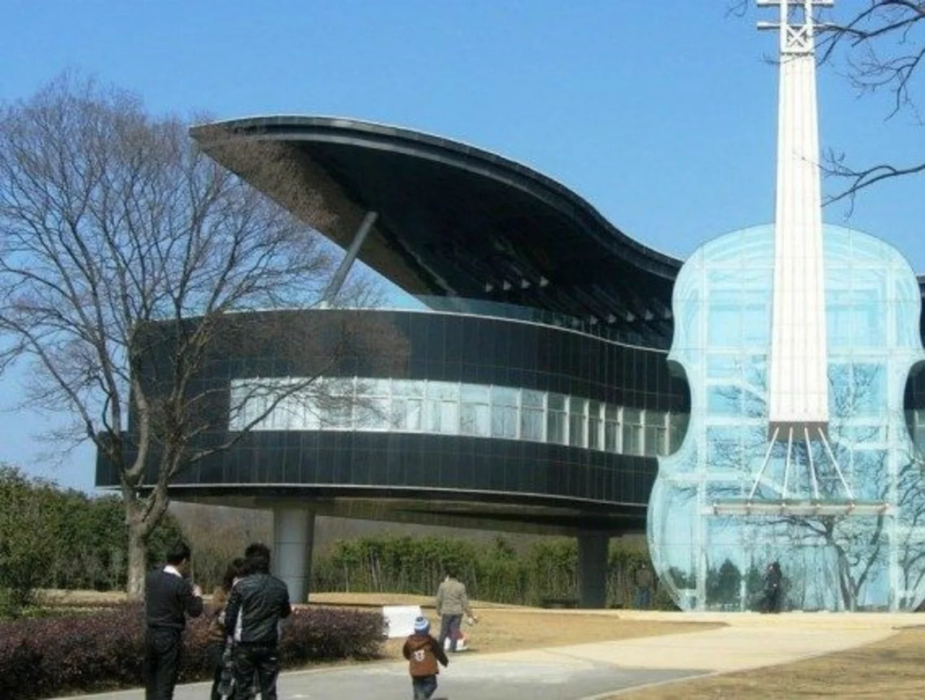 A modern building shaped like a grand piano and a glass violin, with people walking on a path in front of it, surrounded by trees and grass.