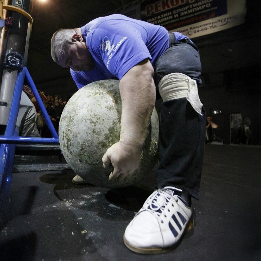 A strongman in a blue shirt and knee wrap is bent over, lifting a large concrete Atlas stone during a strength competition, with a focused expression and an audience watching in the background.