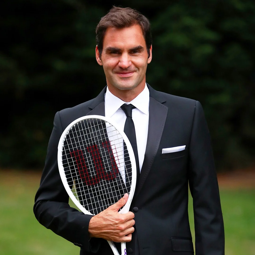 A man in a black suit and tie stands outdoors, smiling and holding a tennis racket upright in front of him. The background is blurred greenery.