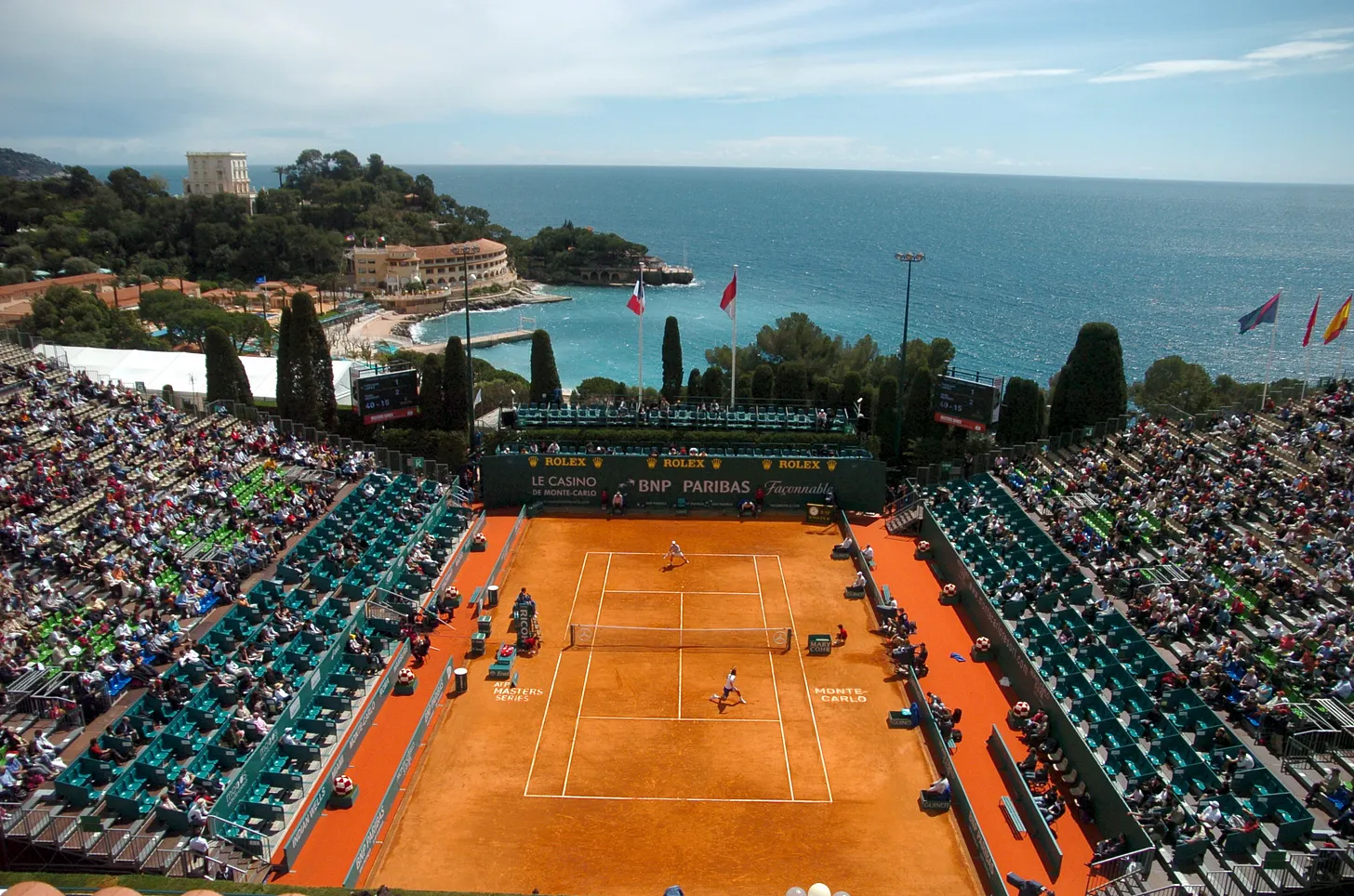 Aerial view of a clay tennis court surrounded by spectators, with the sea and coastal buildings in the background under a partly cloudy sky.