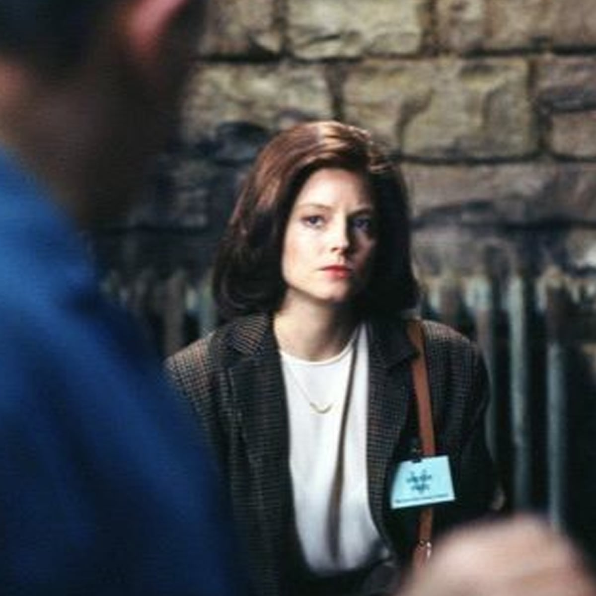 A woman with brown hair in a blazer and white shirt sits in front of a stone wall, wearing a name badge and looking serious. The blurred figure of another person is in the foreground.