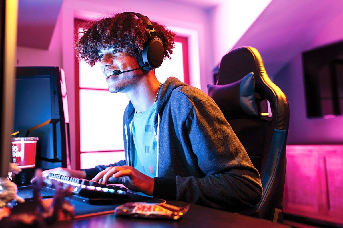 A young man with curly hair wearing a headset and hoodie enthusiastically plays a computer game at a desk, illuminated by colorful purple and pink lighting. Snacks and a drink are visible on the desk.